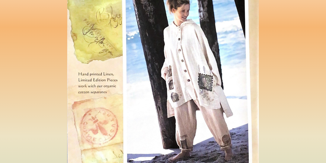 A woman standing on a beach wearing a natural Hooded Cardigan, next to a Boardwalk pillar. A close-up of a paper mailing stamp is on the Cardigan representing the Travel Theme from 1998 Resort collection