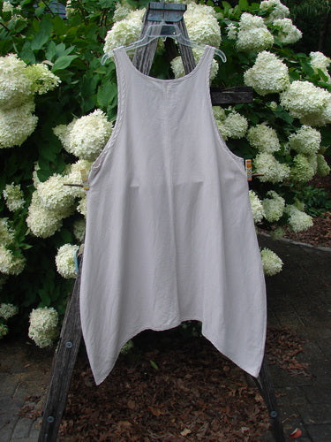 Barclay Hemp Silk Everyway Pinafore in unpainted palest peach, size 1, hangs from a wooden ladder-style stand. The sleeveless garment has a soft, draped A-line shape with a crinkled hemp-silk texture, set against dense white hydrangeas and dark green foliage.