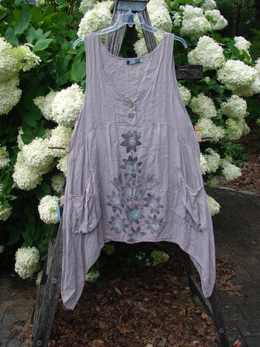 Bluefishfinder.com Barclay Linen Tuesday's Dress Majestic Petal Mallow Size 0 in soft mallow gray linen, hanging from a wooden stand before white hydrangeas. The sleeveless dress has a gathered V-neck, button front, side pockets, and a faded floral print down the front.
