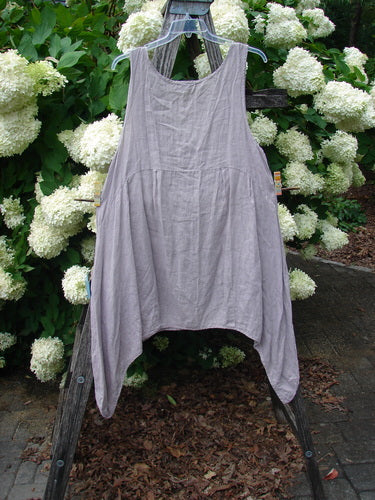 Bluefishfinder.com Barclay Linen Tuesday's Dress Majestic Petal Mallow, a size 0 sleeveless linen dress in soft mallow gray with a square neckline, gathered waist, and draped handkerchief hem. It hangs on a wooden rack before white hydrangeas and dark greenery.
