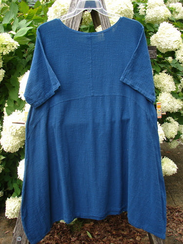 Barclay NWT Textured Gauze Chaya Tunic in Window Meditation Indigo, size 1, with a loose A-line silhouette, short draped sleeves, and a softly crinkled gauze texture. The indigo fabric hangs from a clear hanger, framed by white flowers and green foliage.