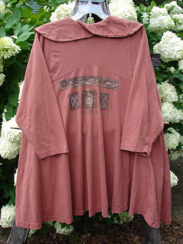 Bluefishfinder.com 1994 Wanderer's Jacket Continuous Garden Monrovian Brick Size 2 in muted brick pink, a loose, cape-like jacket with a rounded sailor collar, wide sleeves, and a gently flared drape. The soft matte fabric shows a centered decorative chest motif, hanging against dense white blossoms and green leaves.