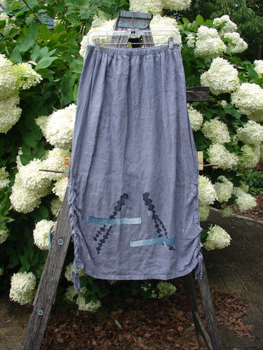 Bluefishfinder.com 1997 Breeze Skirt Sealife Naya size 1 in gray, a long lightweight skirt with a gathered elastic waist and subtle sealife motif near the hem. It hangs on a wooden frame amid white hydrangeas and green foliage, with soft outdoor light emphasizing the woven texture.