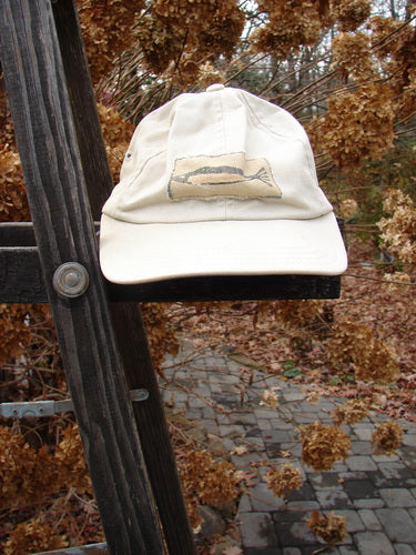 1999 Patched Men's Baseball Cap Single Fish BF Logo Natural OSFA, displayed on a wooden ladder, features a fish patch, adjustable rear strap, and slightly faded brim edges.