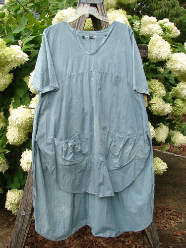 Barclay Percale Key Pocket Hi Low Dress in mottled blue cloud, size 0, with a loose V-neck silhouette, short sleeves, and oversized front pockets. The lightweight woven fabric hangs in soft folds, photographed against white hydrangeas and weathered wood.