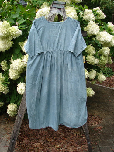 Barclay Percale Key Pocket Hi Low Dress in mottled blue cloud, size 0, hangs from a rustic wooden easel outdoors. The airy percale dress has a gathered waist, short wide sleeves, and a softly draped hi-low silhouette with a matte, lightly textured finish.
