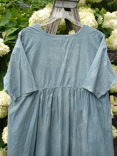 Barclay Percale Key Pocket Hi Low Dress in mottled blue cloud, size 0, with a soft percale texture, short sleeves, a gathered waist, and a loose hi-low silhouette. The dress hangs front-facing, its muted blue fabric set against pale flowers and greenery.