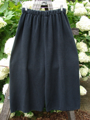 Bluefishfinder.com 1993 Thermal Waffle Wide Leg Crop Pant in unpainted black, size 1, with a textured waffle knit, elastic waistband, and wide cropped legs. The dark fabric hangs from a hanger against leafy white flowers, creating a simple outdoor product display.