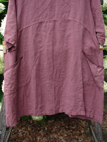 Barclay Summercloth Ditsy Trim Urchin Dress in unpainted loam, size large 0, shown in a muted mauve-brown linen-like fabric with a loose, gently draped silhouette, curved seam details, and short rolled sleeves. The dress hangs front-facing against leafy greenery, with soft natural light emphasizing its textured, relaxed finish.