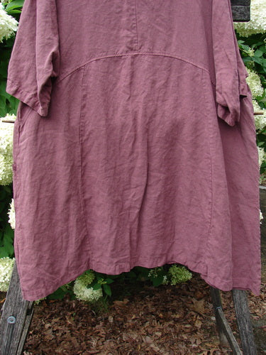 Barclay Summercloth Ditsy Trim Urchin Dress in unpainted loam, size 0, shown in a loose, plum-brown silhouette with short sleeves, a softly curved seam across the back, and a gently flared hem. The textured cloth hangs over weathered wooden supports, with green foliage and pale blooms behind it.