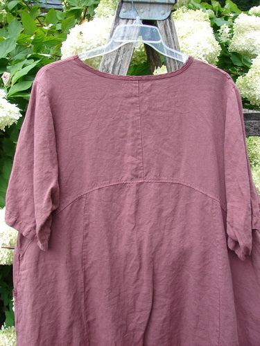 Barclay Summercloth Ditsy Trim Urchin Dress in unpainted loam, size 0, shown as a mauve, crinkled dress with a rounded neckline, short gathered sleeves, and soft vertical and curved seam lines. It hangs on a clear hanger outdoors against green foliage and white flowers.