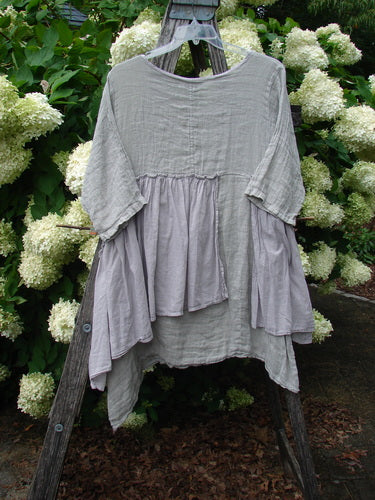 Barclay Gauze Batiste Petal Urchin Flounce Dress in unpainted rice paper, size 1, hangs on a wooden A-frame amid white hydrangeas. The loose gauze batiste dress has a soft gray-beige tone, layered flounced panels, and airy, sheer texture.