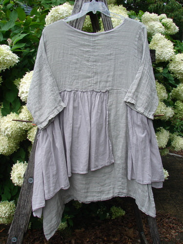 Barclay Gauze Batiste Petal Urchin Flounce Dress in unpainted rice paper, size 1, hangs on a clear hanger with a softly crinkled gauze-batiste texture and layered flounce panels. The pale gray-white dress is shown against dense green foliage and white hydrangea blooms.