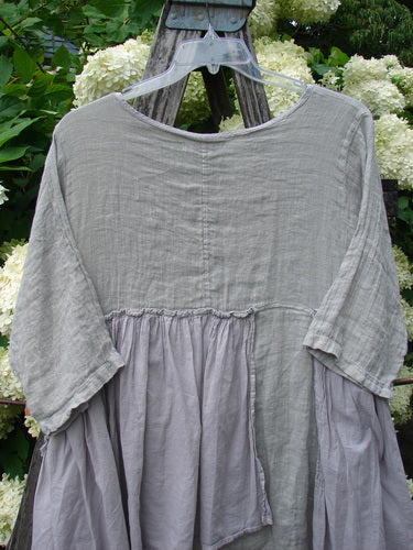 Barclay Gauze Batiste Petal Urchin Flounce Dress in unpainted rice paper, size 1, with a loose, airy silhouette and softly crinkled gauze-batiste texture. The pale gray dress shows a wide rounded neckline, elbow-length sleeves, and gathered flounce panels draping over a rustic hanger outdoors.