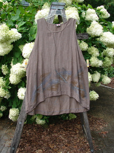 Barclay Hemp Cotton Silk Voile Venetian Pinafore Dragonfly in mocha, size 0, is a sleeveless, loose-cut top with a softly draped A-line shape and sheer, lightweight voile texture. A subtle dragonfly motif appears near the hem, and the garment hangs on a wooden display stand before white hydrangeas and green foliage.