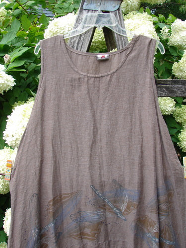 Barclay Hemp Cotton Silk Voile Venetian Pinafore Dragonfly in mocha, size 0, a sleeveless dress with a soft scooped neckline and sheer, crinkled voile texture. Blue dragonfly motifs gather along the lower hem, while the garment hangs on a clear hanger against leafy greenery and white blossoms.