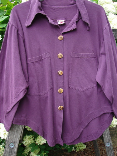 Barclay Wood Button Workshirt in unpainted plum berry, size 0, shown as a purple long-sleeve shirt with a soft collar, five wood buttons, and two chest pockets. The matte fabric hangs loosely against a leafy outdoor backdrop.