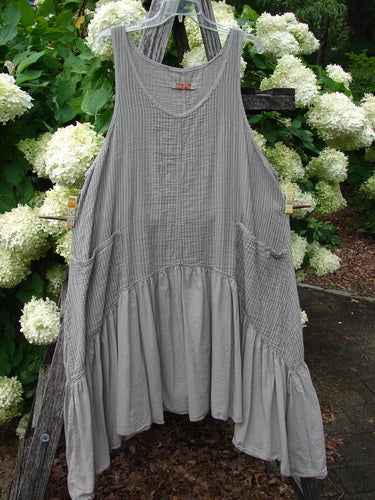 Barclay Gauze Batiste Summer Frock Jumper Stripe Sand Stone Size 1 in sand-stone gray, a sleeveless gauze batiste dress with narrow vertical stripes, a scoop neckline, side pockets, and a gathered, tiered hem. It hangs against leafy white hydrangeas, with soft natural light emphasizing the airy texture.
