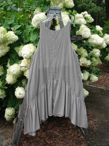 Barclay Gauze Batiste Summer Frock Jumper Stripe Sand Stone Size 1, a sleeveless gray gauze dress with fine vertical stripes, a scoop neckline, and a softly gathered tiered hem. It hangs from a wooden ladder amid white hydrangea blooms, with muted stone and green tones behind it.