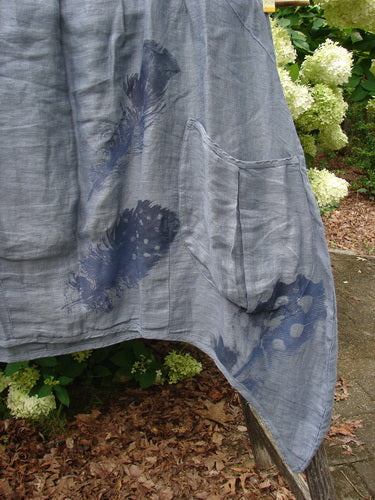 Barclay Linen Tuesday's Dress Guinea Feather Grey Storm size 0 in feather gray, a flowing linen dress with dark feather-like prints and a soft, crinkled texture. The fabric drapes in loose vertical folds, with a curved hem and a visible side pocket against an outdoor garden backdrop.