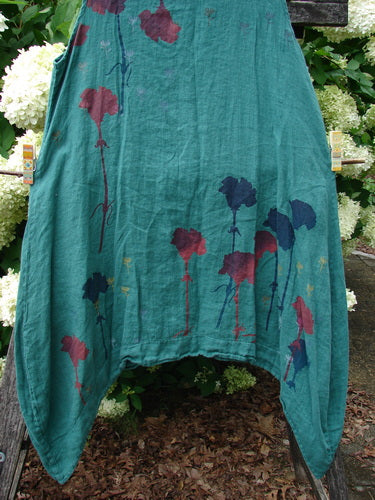 Barclay Linen Round Bottom Pinafore in Cloud Burst Jade, size 0, with a loose linen drape and rounded hem. The teal-green fabric is printed with slender floral stems in burgundy, navy, and yellow, hanging against a leafy garden backdrop.