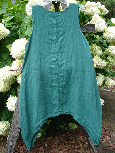 Barclay Linen Round Bottom Pinafore in Cloud Burst Jade, size 0, hangs front-facing in a sleeveless A-line shape with a rounded neckline and pointed hem. The linen has a soft, textured drape, set against leafy white hydrangeas and weathered wood.