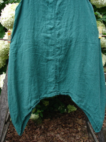 Barclay Linen Round Bottom Pinafore in Cloud Burst Jade, size 0, with a softly draped linen body, center seam, and rounded hem that falls into a pointed front opening. The teal-green fabric hangs against a garden backdrop of white flowers and wood mulch, emphasizing its textured weave and airy shape.