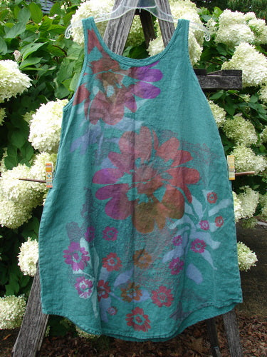 Barclay Linen Round Bottom Pinafore Giant Floral Jade Size 0 in teal jade linen, with a loose sleeveless A-line shape and a rounded hem. Large layered floral prints in burgundy, purple, and pink spread across the front, hanging from a wooden ladder amid white hydrangeas and green foliage.