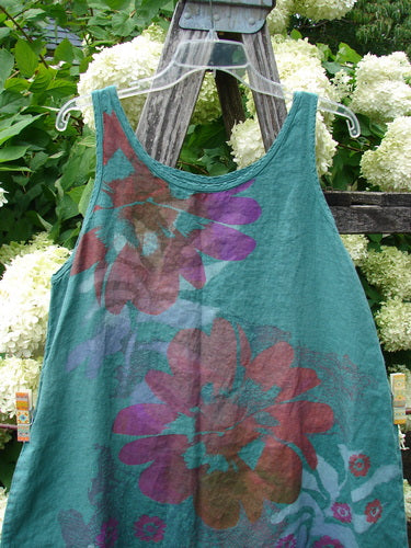 Barclay Linen Round Bottom Pinafore in jade, size 0, with oversized floral prints in burgundy, purple, and teal across a sleeveless, round-neck linen silhouette. It hangs on a clear hanger against white hydrangeas and weathered wood, with soft daylight bringing out the textured weave and layered colors.