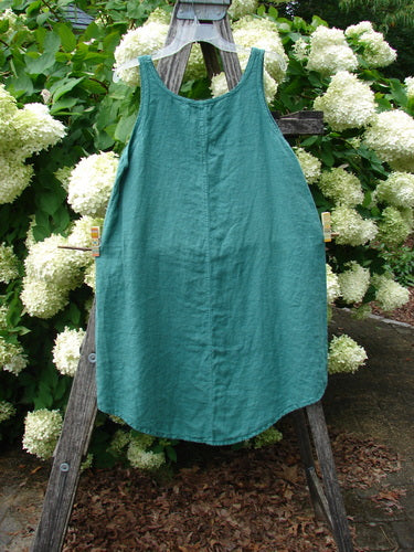 Barclay Linen Round Bottom Pinafore Giant Floral Jade Size 0 in teal-green linen, with a sleeveless scoop neckline and softly rounded hem. It hangs centered on a weathered wooden ladder, framed by dense white hydrangea blooms and dark green foliage.