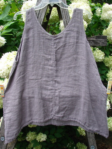 Barclay Linen Altoll Top Wing Stats Rose Quartz Size 0 in muted rose quartz linen, a sleeveless top with a wide V neckline, center seam, and softly flared hem. It hangs against a rustic wooden backdrop amid pale flowers and green foliage.