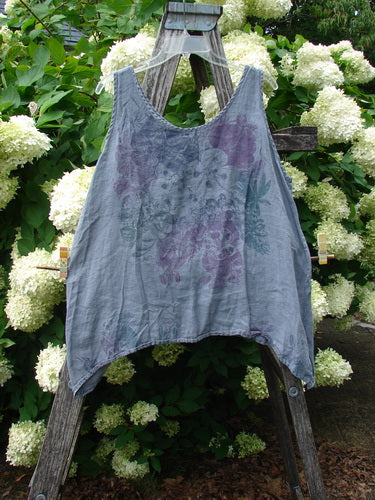 Barclay Linen Altoll Top Pansy Patch in blue dust, size 0, is a sleeveless linen top with a soft V neckline, wide straps, and a loose draped hem. The faded blue-gray fabric carries a painterly purple floral patch, hanging against a rustic wooden ladder amid white hydrangeas.