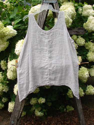 Barclay Linen Altoll Top Floral Victorian Floral Natural Size 0 in light natural linen, a sleeveless swing top with a soft V neckline, wide straps, and an uneven hem. It hangs on a rustic wooden ladder amid white hydrangeas and dense green foliage.