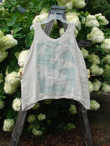 Barclay Linen Altoll Top Blossom Reach Creme Size 0, a sleeveless cream linen top with a loose, draped silhouette and faded blue-green graphic on the front. It hangs from a weathered wooden ladder amid white hydrangeas and dense green foliage.