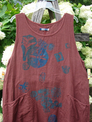 Barclay Summercloth Round Pocket Pinafore Star Flower Rusty Bucket Size 0 in a rusty maroon tone, made from soft, textured fabric with a round neckline and wide shoulder straps. Blue star-and-flower prints spread across the front, and large curved patch pockets sit low on the skirt.