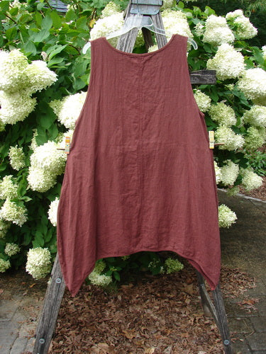 Barclay Summercloth Round Pocket Pinafore Star Flower in rusty bucket, size 0, a sleeveless linen dress with a rounded neckline and softly draped A-line shape. It hangs from a weathered wooden ladder, its muted rust color set against dense green foliage and pale white blossoms.