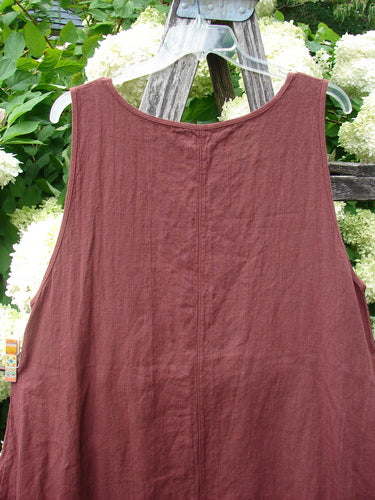 Barclay Summercloth Round Pocket Pinafore Star Flower Rusty Bucket Size 0 in a muted rusty red, shown as a sleeveless pinafore with a rounded neckline and soft crinkled texture. It hangs from a clear hanger against white flowers and green foliage, with a weathered wooden ladder behind it.