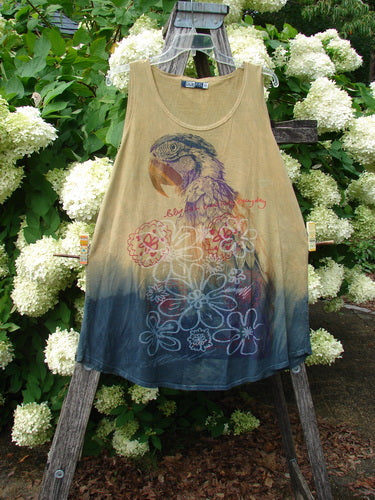 Barclay Round Bottom Tunic Tank Top Parrot Mottled Golden Sunset Size 0 in a sleeveless A-line silhouette, with a faded golden-tan body, dark blue dipped hem, and layered parrot and floral graphics. The soft, worn fabric hangs from a rustic wooden stand amid white hydrangeas and green foliage.