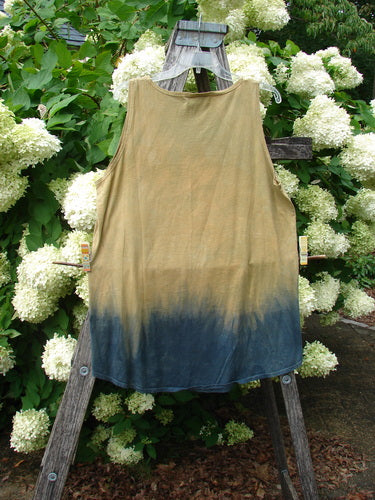 Barclay Round Bottom Tunic Tank Top in Parrot Mottled Golden Sunset, size 0, hangs from a weathered wooden ladder in front of dense white hydrangeas. The sleeveless top has a soft, draped shape and a tan body that fades into mottled blue at the hem, with a muted, natural finish.