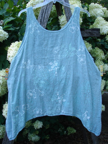 Barclay Linen Altoll Top Falling Floral Sage Size 2, a loose sleeveless sage-blue linen top with a softly flared hem and faint falling floral pattern. It hangs on a hanger, showing a light, airy weave against dense green foliage and white flowers.
