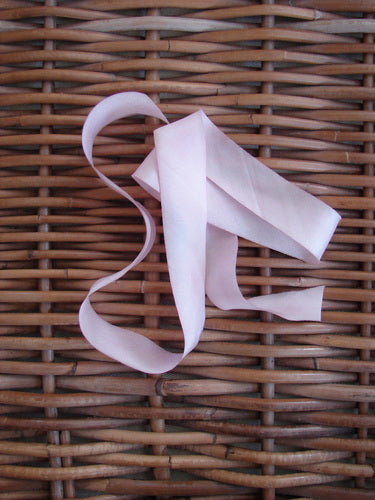 Blue Fish Hand Dyed Silk Ribbon in Victorian Rose 108 x 1, delicately laid on a woven wicker surface, showcasing its light and flowing, hand-dyed texture with unique color variations.