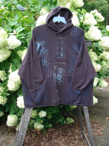 Barclay Cotton Lycra Dolman Roo Pocket Hoodie in Pansy Rose Deep Bark, a loose dark brown pullover with a draped hood, oversized dolman sleeves, and a large front roo pocket. The soft cotton-lycra knit shows blue printed accents and hangs from a hanger outdoors.