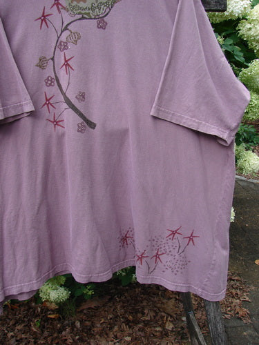 Barclay Short Sleeved Studio Tee Tree Star Laurel Size 3 in dusty lavender cotton, a loose short-sleeve T-shirt with a soft drape and faded graphic print of tree branches, stars, and laurel-like details. The hem hangs unevenly over leafy outdoor greenery.
