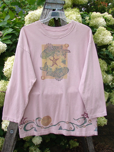 1993 Long Sleeved Tee Ocean Folly Ask Pink OSFA in soft pink cotton, shown with a loose crewneck silhouette and dropped shoulders. The front features a centered ocean-inspired graphic, while the hem carries a flowing decorative border, all hanging on a clear hanger against leafy white blooms.