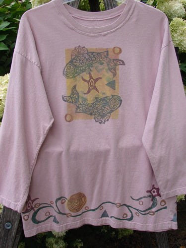1993 Long Sleeved Tee Ocean Folly Ask Pink OSFA in soft pink cotton, with a loose long-sleeve cut and a faded central graphic framed by a decorative border. The hem carries a flowing vine-like print in muted green and burgundy, set against leafy outdoor greenery.