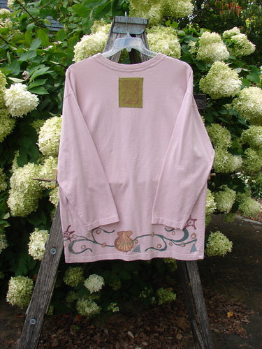 1993 Long Sleeved Tee Ocean Folly Ask Pink OSFA in soft pink, a long-sleeved tee with a relaxed drape, square olive patch at the upper back, and ornate scroll-and-shell hem print. It hangs on a wooden ladder amid white hydrangeas.