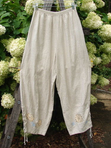 1997 Skipper Pant Rolling Sealife Natural Size 1 in natural beige linen, with a loose, tapered silhouette and gathered elastic waist. The pants hang front-facing against dense green hydrangea blooms, showing soft texture, a central seam, and small sealife motifs near the hems.