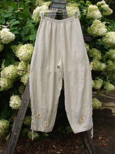1997 Skipper Pant Rolling Sealife Natural Size 1 in natural beige, loose-legged pants with a soft, textured fabric and small sea-life motifs near the hems, hanging from a wooden stand against dense green hydrangeas.