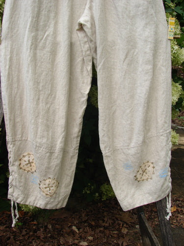 1997 Skipper Pant Rolling Sealife Natural Size 1 in natural linen-toned fabric, shown as loose pants with wide legs, a soft wrinkled texture, and small sealife motifs near the hem. The cropped view centers the lower legs against leafy greenery.