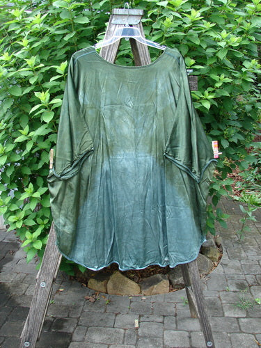 Barclay Viscose Vitreous High Low Tunic Botanicals Mottled Forest Sun OSFA displayed on a wooden rack, showcasing its rounded neckline, varying hem, and three-quarter dolman sleeves with botanical-themed exterior stitchery.